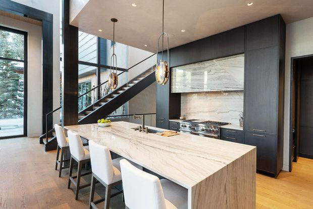 A sophisticated open-plan kitchen with a large marble-look island and waterfall edge, seating for several people, dark full-height cabinetry, integrated appliances, and a dramatic stone feature backsplash. Pendant lights hang above the island, while a sculptural stair and extensive glazing create a bright, double-height feel.