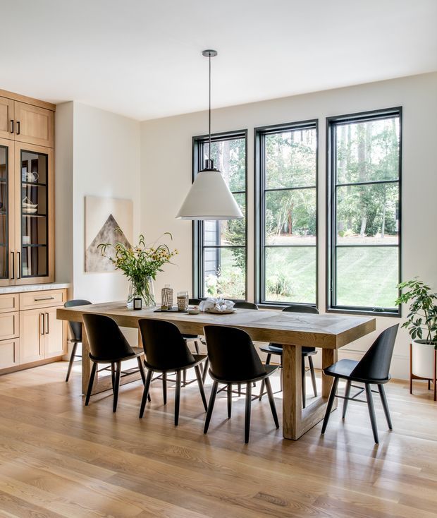 Bright, refined dining area with a long solid-wood dining table and six black upholstered chairs, positioned beside three tall black-framed windows overlooking greenery. A large white pendant light hangs above the table. The room includes light timber flooring, warm beige walls, and built-in timber cabinetry with glass-front display storage along the left wall, complemented by indoor plants and understated artwork.