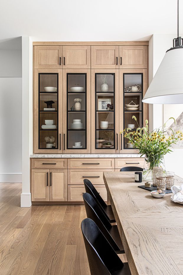 Bright, refined kitchen and dining area featuring a full-height light timber built-in cabinet wall with glazed display doors, drawers, and lower cupboards. A pale stone benchtop separates the cabinetry from the large light timber dining island, accompanied by black upholstered stools. The room has light timber flooring, white walls, and a large pendant light, with styling objects and flowers adding warmth.