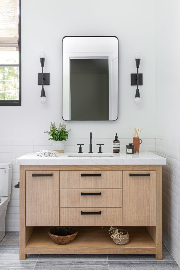 Bright, compact contemporary bathroom vanity area with a light timber floating-style cabinet, white stone benchtop, integrated basin, matte black tapware and wall sconces. White horizontal subway tiles cover the lower walls, while a large rounded-rectangle mirror is centred above the vanity. Grey wood-look floor tiles and a partially visible toilet complete the room.
