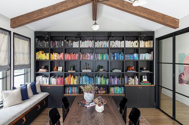 A bright, refined home library and dining area with a full-height dark built-in bookcase covering the back wall, filled with books and decorative objects. A rustic timber dining table with black chairs sits in the foreground, while a built-in window seat with cushions occupies the left side. Exposed timber ceiling beams, white walls, black-framed glazing and doors, and warm timber flooring define the room.
