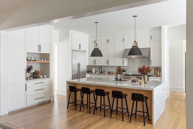 Bright, spacious contemporary kitchen with extensive white shaker-style cabinetry, integrated appliances, a large marble-look island with seating for five, and two black pendant lights. Warm timber flooring contrasts with the light cabinetry, while a mottled stone backsplash and island waterfall sides add texture. The kitchen opens to an adjacent area through a wide opening.