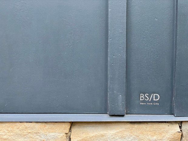 Close-up detail of a dark grey-blue painted exterior panel or cladding with vertical seams, resting above rough pale natural stone masonry. A small “BS/D New York City” mark is printed on the panel. The image focuses on exterior building materials and their junction.