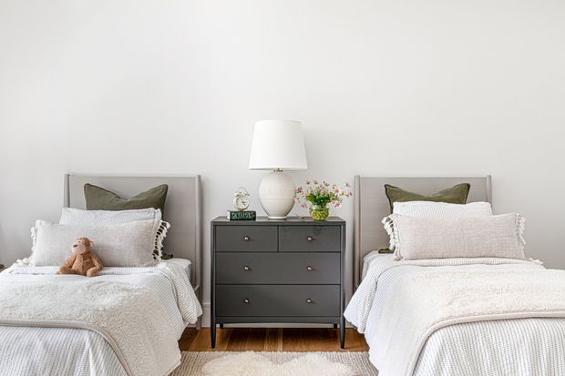 A bright, symmetrical bedroom with two single beds separated by a dark chest of drawers. The beds are dressed in layered white and light-grey bedding with olive-grey accent cushions, while a table lamp, small clock, greenery and decorative objects sit on the central dresser. Timber flooring and a pale neutral wall create a clean, calm setting.