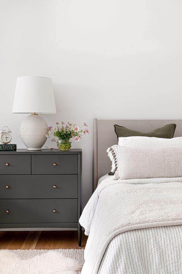 A softly styled bedroom corner with a dark charcoal six-drawer dresser beside a bed. The bed has a pale taupe upholstered headboard, layered white and cream textured bedding, and an olive green accent cushion. A ceramic table lamp, small floral arrangement, alarm clock, and decorative objects sit on the dresser. The room has light neutral walls, warm timber flooring, and a plush cream rug, creating a calm, cozy, refined atmosphere.