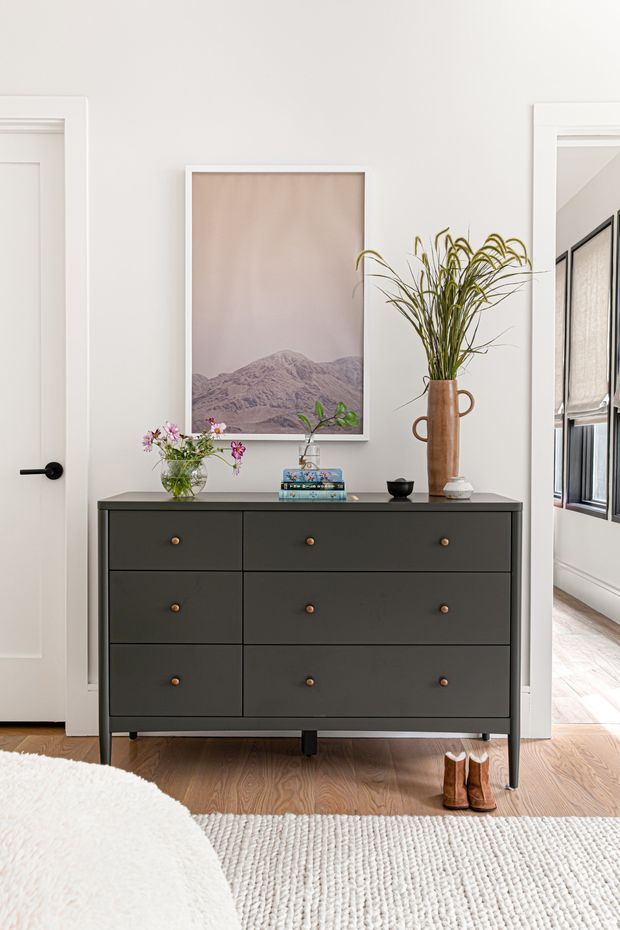 A styled hallway or entry area with a dark charcoal six-drawer console against a white wall. The console is topped with flowers, books, a small bowl, and a tall ceramic vase with greenery. A large framed landscape print hangs above, with white-trimmed doorways on either side. Timber flooring, a light textured rug, and boots near the console add warmth. A bright adjoining room with windows is visible through the doorway on the right.