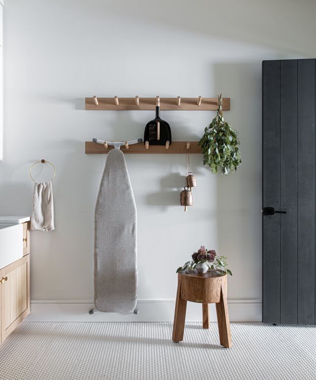 A bright, calm entry area with pale grey walls, a textured light carpet, and simple timber wall-mounted coat racks holding clothing and accessories. A narrow console stool with a potted floral arrangement sits beside a tall dark timber door. A small timber cabinet is partially visible at left, and a circular wall-mounted towel or garment holder adds a subtle decorative detail.