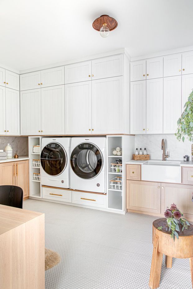 Bright, spacious laundry room with extensive white shaker-style cabinetry, integrated open shelving, front-loading washer and dryer, a large farmhouse-style utility sink, and timber-toned lower cabinetry. A pale patterned floor, marble-look backsplash, brass hardware, timber work surfaces, and a copper-toned ceiling light create a refined, warm traditional interior. A small dining or work surface and plant styling are visible in the foreground.