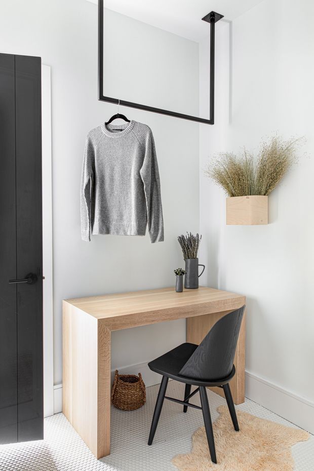 A bright, compact entryway or dressing nook with white walls, a black door, a suspended black metal hanging rail displaying a grey knit sweater, and a light timber console desk. A black chair, woven basket, small vases, dried grasses, and a textured rug add functional and decorative detail. The space has a clean, minimalist contemporary aesthetic with strong black-and-white contrast and warm timber accents.