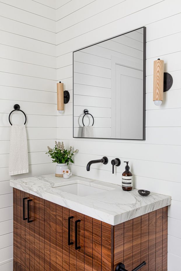 Bright, compact bathroom vanity with white horizontal wall panelling, a large black-framed rectangular mirror, black wall-mounted tapware and sconces, and a warm timber vanity topped with a white marble-look benchtop and integrated basin. A towel ring, hand towel, soap dispenser and small floral arrangement complete the styled space.