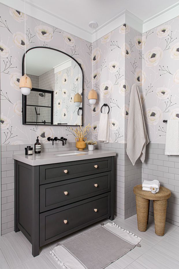 A bright, compact bathroom with a dark charcoal vanity featuring three drawers, a pale stone benchtop, wall-mounted black tapware, and a rectangular undermount basin. The walls combine grey subway tile wainscoting with light floral wallpaper above. A large arched black-framed mirror, warm pendant lights, woven baskets, towels, and a neutral fringed rug create a playful yet refined look.