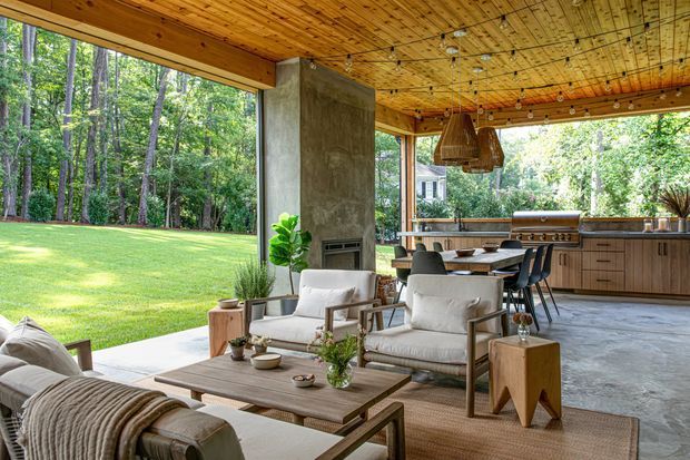 A spacious, open-sided covered outdoor living area seamlessly connected to a modern kitchen. The space features a timber-lined ceiling with exposed wiring and pendant lights, a concrete structural column and floor, a built-in linear fireplace, a timber kitchen with island seating, and a relaxed seating arrangement overlooking a landscaped wooded garden.