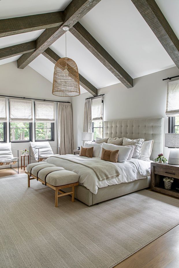 A bright, spacious bedroom with a large upholstered bed, layered white and taupe bedding, matching upholstered bench, bedside tables, and lounge chairs by the windows. The room features white walls, large windows with Roman blinds and curtains, a neutral carpet, and a vaulted ceiling with prominent rustic exposed timber beams. A woven pendant light provides a natural textural accent.