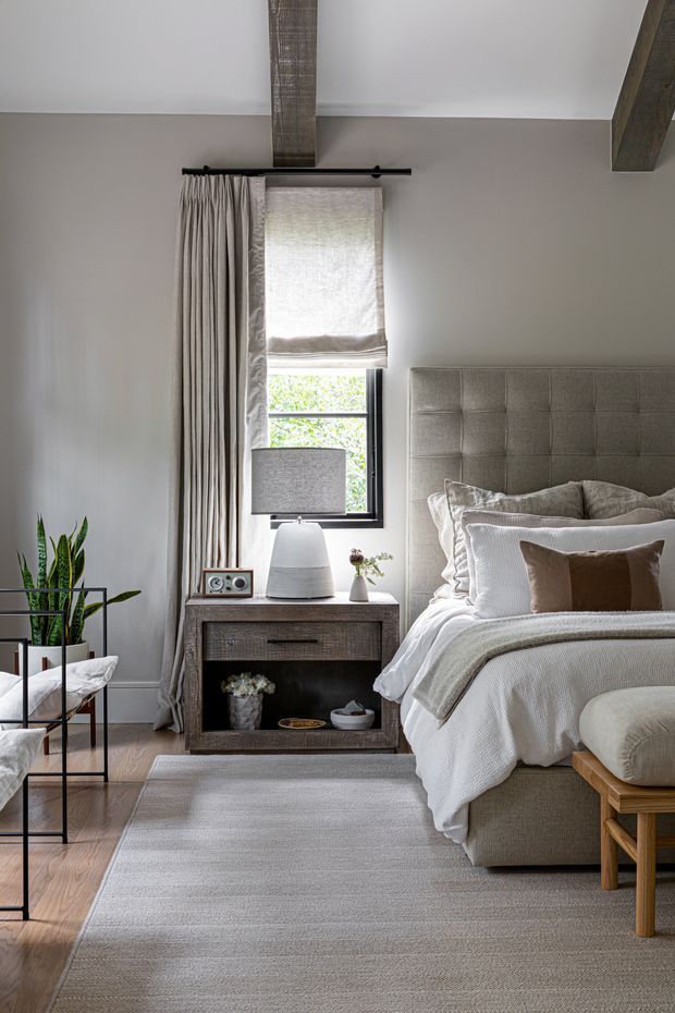 A refined, softly lit bedroom with a large upholstered tufted headboard, layered white and taupe bedding, a wooden bedside table, table lamp, framed window with Roman shade and full-length curtains, exposed timber ceiling beams, indoor plants, and a neutral textured rug. The room has a warm, relaxed contemporary-traditional feel.