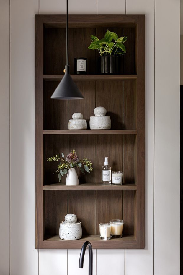 A close-up interior view of a dark walnut-toned built-in recessed shelving unit set into vertically panelled white walls. The shelves display plants, ceramic vessels, candles, bottles and decorative objects, with a matte black pendant light hanging in front and a black curved tap visible at the bottom, suggesting the joinery is above a kitchen or wet-area counter. The composition is warm, refined and contemporary, with strong vertical lines and dark timber contrast.