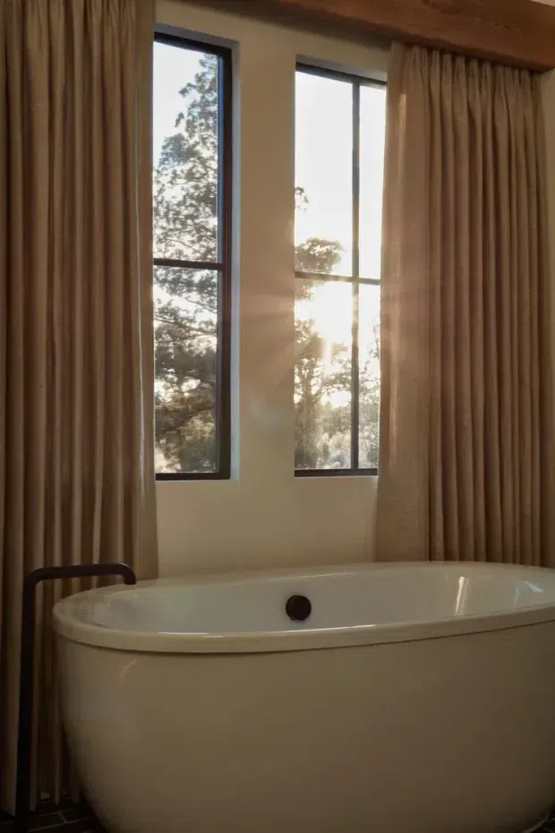A freestanding white bathtub occupies the foreground of a warm, softly lit bathroom. Tall narrow dark-framed windows look onto trees, with full-height beige pleated curtains on either side. A dark floor-mounted tub filler is visible beside the bath, and a timber-toned ceiling or beam adds warmth.