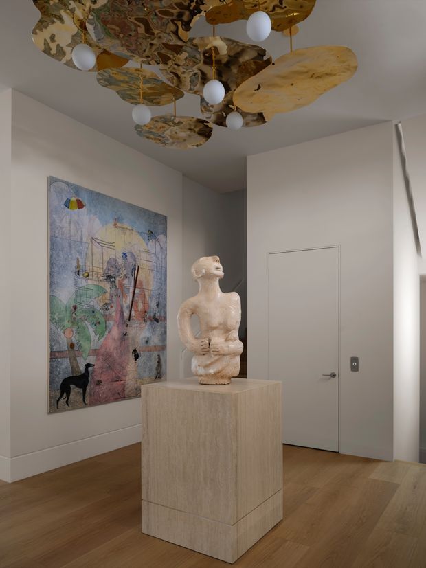 A refined residential interior vignette showing a sculptural artwork on a stone-clad pedestal in a softly lit hallway or entry space. The room has warm timber flooring, pale neutral walls, a flush concealed door, contemporary artwork, and an expressive sculptural ceiling installation with organic patterned panels and spherical pendant lights.