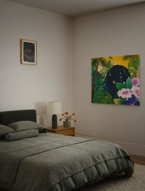 A cozy bedroom with a low-profile upholstered bed dressed in layered grey bedding, a timber bedside table, table lamp, and small floral arrangement. Neutral warm-grey walls and carpet create a calm backdrop, while colourful botanical artwork provides a vivid accent on the main wall.