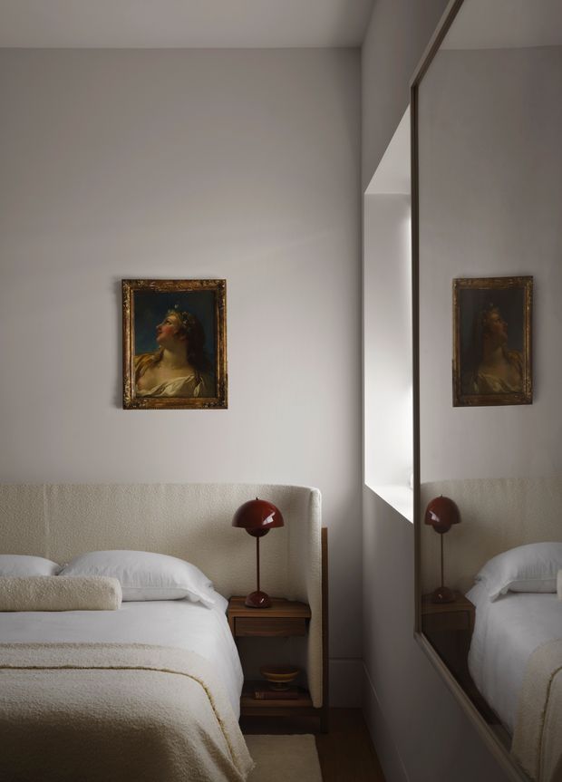 A compact, softly lit bedroom with a white textured upholstered headboard, white bedding, a warm wood bedside table, and a dark red dome-shaped table lamp. A framed classical portrait hangs above the bed, while a full-height mirror on the right reflects the bed and artwork. The room has a calm, refined, warm-neutral interior with understated decorative detailing.