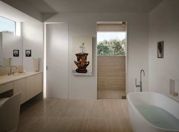 Contemporary minimalist bathroom with a freestanding white bathtub in the foreground, a floating timber-toned vanity with integrated basin and wall mirror to the left, and a frameless glass shower enclosure at the rear. Neutral plaster walls, warm wood-look flooring, restrained artwork, and soft natural light create a calm, refined atmosphere.