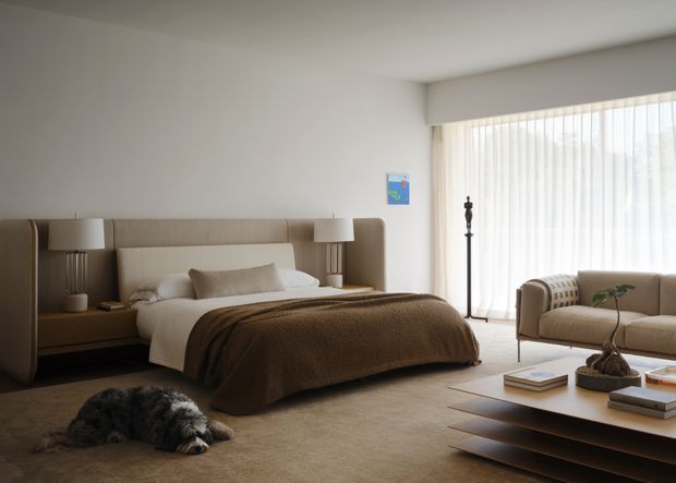 A calm, warm contemporary bedroom with a large upholstered bed and integrated padded headboard spanning the wall, built-in bedside niches and lamps, neutral bedding, and a soft brown textured bedcover. Floor-to-ceiling windows with sheer vertical blinds provide abundant diffused daylight. A low layered coffee table, upholstered lounge chair, decorative floor lamp, books, and a small dog complete the refined residential interior.