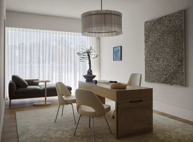 A refined, softly lit open-plan living and dining area with a compact timber dining table or desk, upholstered cream dining chairs, a dark sofa, sheer floor-to-ceiling curtains, and a large textured wall artwork. A sculptural pendant light hangs above the table, while a neutral patterned rug and pale walls create a calm, understated interior.