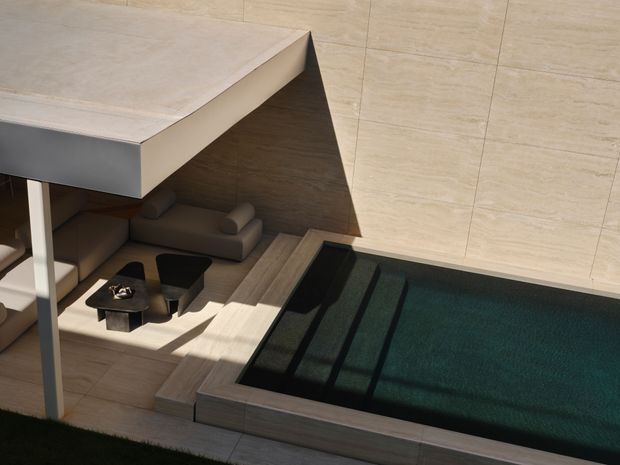A high-angle view of a contemporary outdoor courtyard featuring a rectangular swimming pool with submerged steps, surrounded by pale stone or porcelain paving. A partially covered lounge area sits beside the pool, with built-in seating, a low coffee table, and dark outdoor chairs. The space uses clean geometric forms, warm neutral finishes, and strong sunlight and shadow.