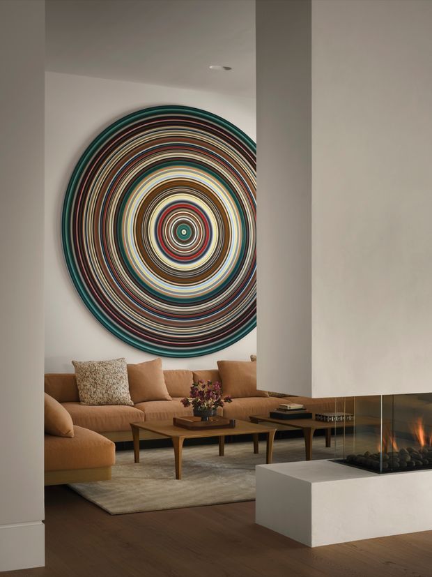 A warm, refined living room viewed through a partial opening, featuring a large circular multicoloured concentric wall artwork above a tan sectional sofa, low wooden coffee tables, a neutral rug, and a contemporary built-in glass-enclosed fireplace in the foreground. White plaster walls, dark timber flooring, and restrained recessed lighting create a sophisticated contemporary setting.