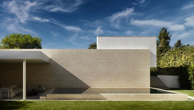 A minimalist modern residential exterior featuring strong rectilinear volumes, a large pale beige stone or concrete-clad wall, a projecting flat roof, and a recessed shaded outdoor lounge. A long narrow reflecting pool or water feature runs along the base of the facade, with manicured lawn and hedging surrounding the house under a bright blue sky.