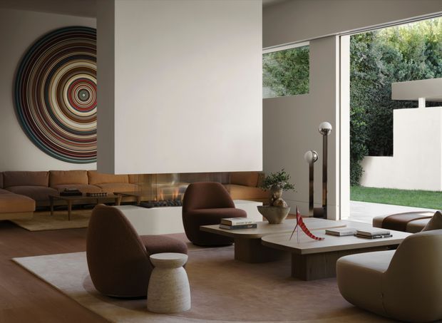 Sophisticated contemporary living room with a warm neutral palette, sculptural brown lounge chairs and sofa, layered low coffee tables, a large suspended rectangular fireplace or feature element, and floor-to-ceiling glazing opening to a landscaped courtyard. A bold circular abstract artwork provides a colourful focal point on the wall.