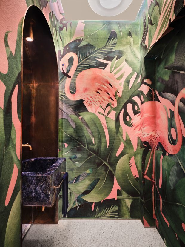 A compact, highly decorative bathroom or powder room with floor-to-ceiling tropical flamingo wallpaper in deep green, coral pink, black, and muted lavender tones. A tall arched niche or shower enclosure with dark bronze-toned metal lining sits on the left, next to a small dark marbled vessel basin and bronze tap. The floor is light grey terrazzo, and a round ceiling light is visible above. The space has a bold, playful, immersive character.
