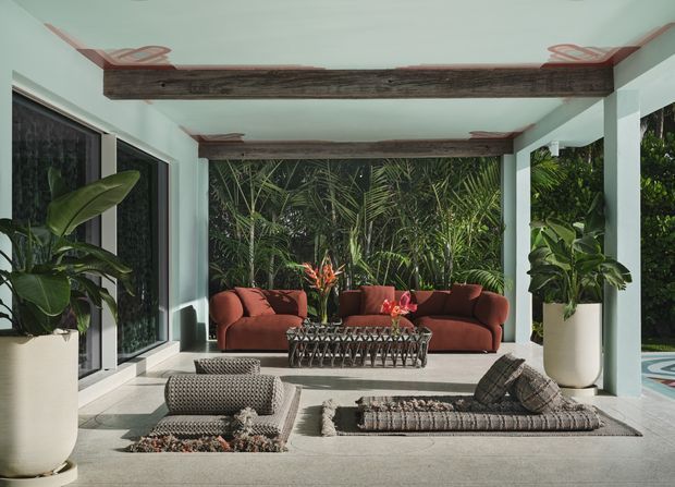 A covered outdoor lounge or sunroom with pale mint-painted walls and columns, exposed dark timber ceiling beams, large dark-framed glazed openings, and patterned concrete or stone flooring. The space is furnished with rust-coloured sofas, low woven lounge seating, a sculptural coffee table, and large potted tropical plants, opening onto dense greenery and a glimpse of a pool.