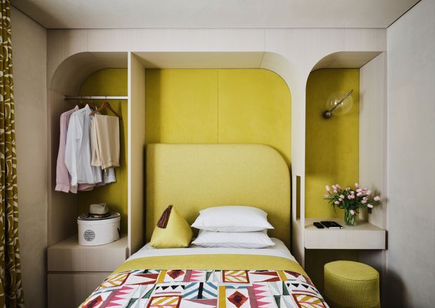 Compact bedroom with a built-in wall of arched niches surrounding the bed. The cabinetry and wall surfaces are light beige, with a vivid mustard-yellow upholstered bed surround and matching niche backing. Open shelving displays hanging clothes and accessories, while integrated bedside storage, a small wall light, and a textured round stool complete the space. A colourful geometric bedspread adds a playful accent.