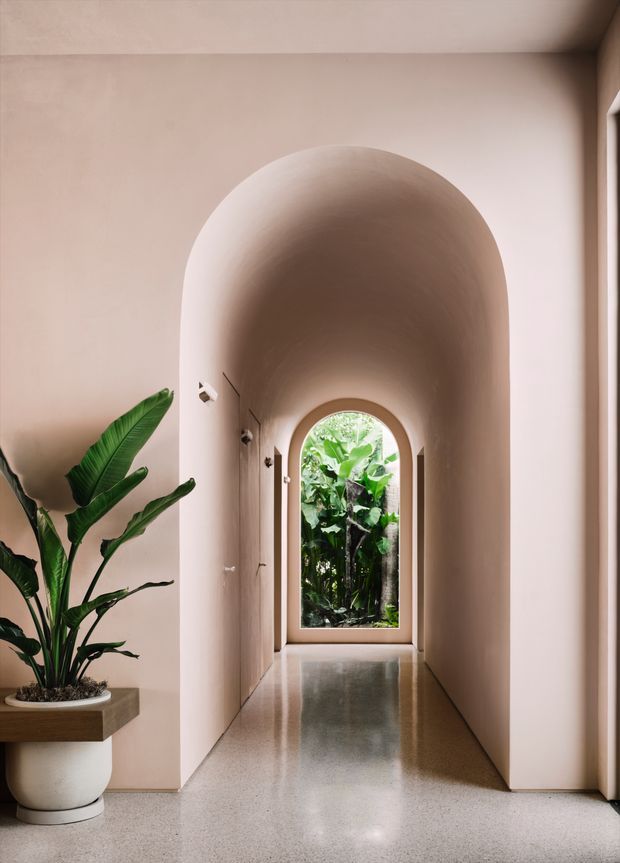 A softly lit residential hallway with a tall, rounded archway leading to a smaller arched glazed door or window overlooking lush greenery. The corridor has smooth warm beige plaster walls, a polished pale terrazzo-like floor, recessed or minimal wall lighting, and a large potted tropical plant on a built-in ledge. The space feels calm, sculptural, and refined, with strong curved architectural detailing.