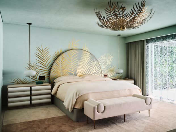 A stylish bedroom with pale green walls, a large upholstered bed, a blush-toned bench at the foot, and matching low dressers beside the bed. A dramatic brass-toned botanical chandelier hangs overhead, while a large circular headboard or wall feature is decorated with layered palm-leaf motifs. Floor-to-ceiling glazing with sheer curtains brings in abundant natural light. The room has a refined, softly glamorous, botanical-inspired aesthetic.