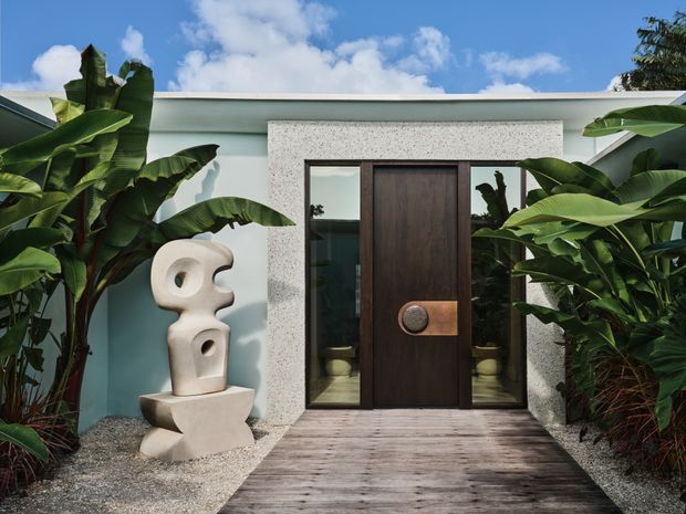 A contemporary single-storey building entrance framed by lush tropical banana plants. A dark timber pivot door with narrow glazed sidelights is set within a light speckled stone or concrete surround, opening onto a timber boardwalk. A pale sculptural figure stands on a pedestal beside the entry, with gravel landscaping and a bright blue sky beyond.
