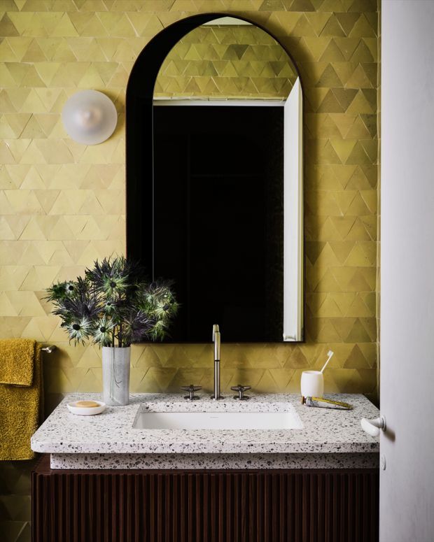 A richly styled bathroom vanity with a white speckled stone benchtop, integrated rectangular basin, dark timber fluted cabinetry, brass-toned fixtures, and a dramatic black arched mirror. The walls are clad in a geometric textured gold-beige tile, with a spherical wall light, mustard towel, and decorative vase adding warmth and character.