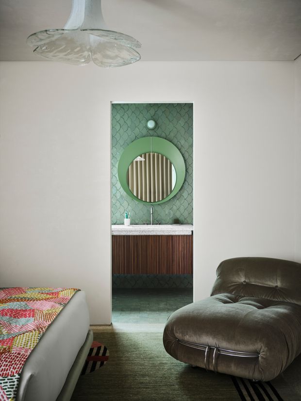 A refined interior view looking from a living room into a compact bathroom or powder room. The bathroom features a green scallop-pattern tiled feature wall, a circular green-framed mirror, fluted timber vanity cabinetry, and a pale stone benchtop. The foreground living area contains a plush dark leather lounge chair, a light upholstered sofa, a patterned textile, and a sculptural glass ceiling light. The palette is muted green, warm timber, neutral walls, and dark leather, with an eclectic retro-luxe character.