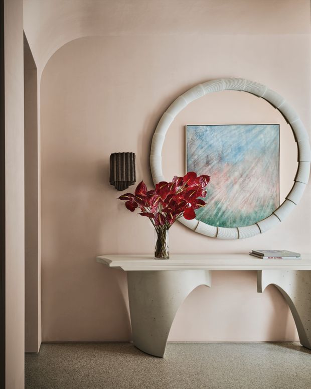 A refined interior vignette featuring a sculptural white console table against a blush-pink plaster wall. The console has curved, architectural supports and a pale stone-like top, with a tall vase of burgundy foliage. Above it hangs a large round mirror with a ribbed pale frame, partially overlapping an abstract blue, pink and green artwork. A dark wall sconce, small books, and a textured grey terrazzo-like floor complete the softly lit setting.