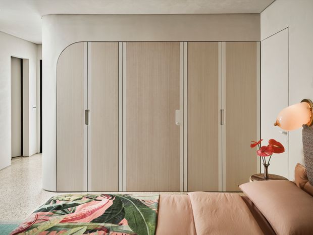 A contemporary bedroom interior featuring a full-width built-in wardrobe with pale timber doors, slim dark vertical detailing, and a rounded end panel. The room has a warm neutral palette, a textured plaster ceiling, terrazzo-style flooring, and a soft pink upholstered bed with a bold botanical bedspread. A sculptural orange bedside lamp and red anthurium arrangement add colourful accents. An adjoining hallway or entry area is visible to the left.