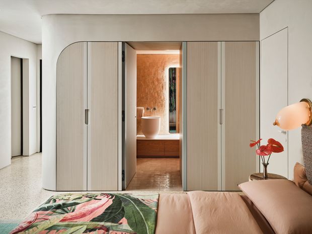 A refined bedroom with a soft neutral palette and built-in full-height cabinetry forming a concealed opening to a warm-toned bathroom. The room features a low upholstered bed with blush bedding, a bold botanical-patterned throw, terrazzo-style flooring, curved cabinetry detailing, and a sculptural wall light. The bathroom beyond includes a freestanding vessel basin, timber-toned joinery, and a textured terracotta feature wall.