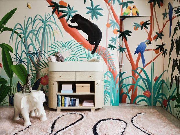 A playful, richly decorated children's room with a full-wall tropical jungle mural featuring oversized foliage, colorful birds, and a black monkey. A rounded light-wood storage cabinet sits against the mural, with books and toys in open shelving. The room includes a white elephant-shaped stool or side table, a spotted giraffe toy, and a soft cream rug with bold black organic lines. The overall look is whimsical, colorful, and nature-inspired.