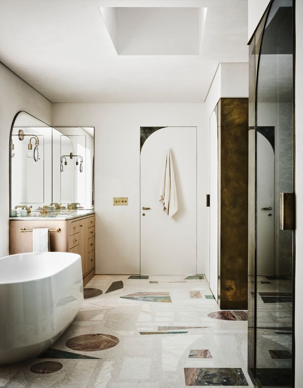 A refined contemporary bathroom with a freestanding white bathtub, glass-enclosed shower, wall-mounted towel, mirrored vanity, brass fixtures, and timber-toned cabinetry. The floor is a decorative pale stone or terrazzo-like surface with inset multicoloured geometric and circular stone details. A large ceiling opening or skylight brings light into the room, while dark metallic and bronze-toned shower framing adds contrast.