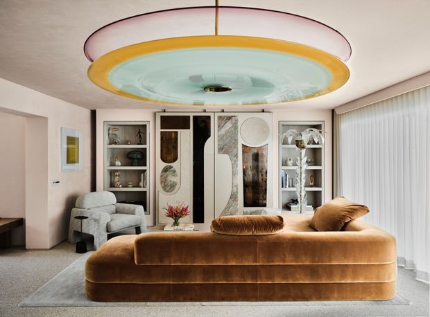 A sophisticated living room with a large sculptural circular pendant light, a plush caramel-brown curved sofa, and a coordinated built-in display and storage wall. The room features pale textured walls, soft carpet flooring, floor-to-ceiling sheer curtains, and decorative marble-like and artistic panels integrated into the joinery. The overall composition is polished, warm, and highly decorative with strong Art Deco-inspired geometric and rounded forms.