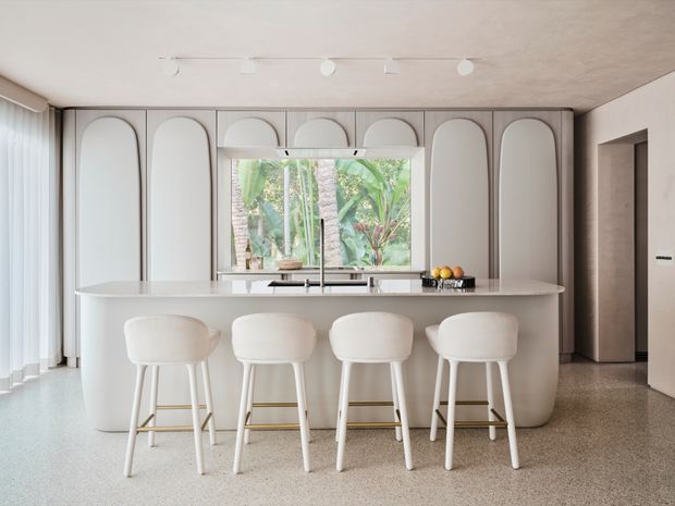 A bright, refined contemporary kitchen viewed from the living area, featuring a large curved island with four white upholstered bar stools, integrated white cabinetry with repeated arched panel detailing, a compact sink beneath a window, and pale terrazzo-style flooring. Full-height glazing at left and tropical greenery visible through the window add an airy connection to the outdoors. A minimalist ceiling track light and soft neutral finishes complete the space.