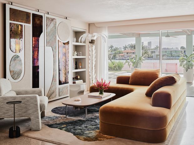 Bright, refined living room with a large rounded brown sectional sofa, sculptural coffee table, textured rug, boucle lounge chair, built-in open shelving, and a dramatic floor-to-ceiling decorative wall installation. Full-height glazing opens to a landscaped outdoor setting with water visible beyond, while soft white walls and curtains create an airy atmosphere.