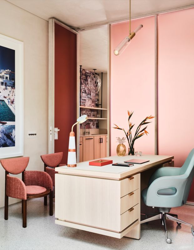 A stylish compact home office with a pale timber desk featuring integrated drawers and a light benchtop, positioned in front of pink wall panels. The room includes a pale blue swivel office chair, two dusty rose upholstered guest chairs, a sculptural desk lamp, pendant light, open shelving with books and objects, framed artwork, and decorative flowers. The palette is warm pastel pink, coral, pale timber, cream, and muted blue, with a polished terrazzo-like floor.