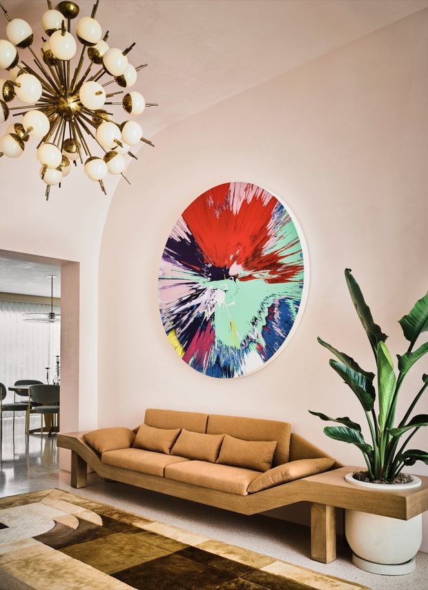 A stylish living room with a warm blush-beige plastered wall, a sculptural mustard-brown upholstered sofa on a timber base, and a large colourful abstract circular artwork. A dramatic brass-and-globe chandelier hangs overhead, while a tall potted tropical plant sits in a built-in timber planter beside the sofa. A patterned area rug defines the seating zone, and an open passage reveals a contemporary dining area beyond.