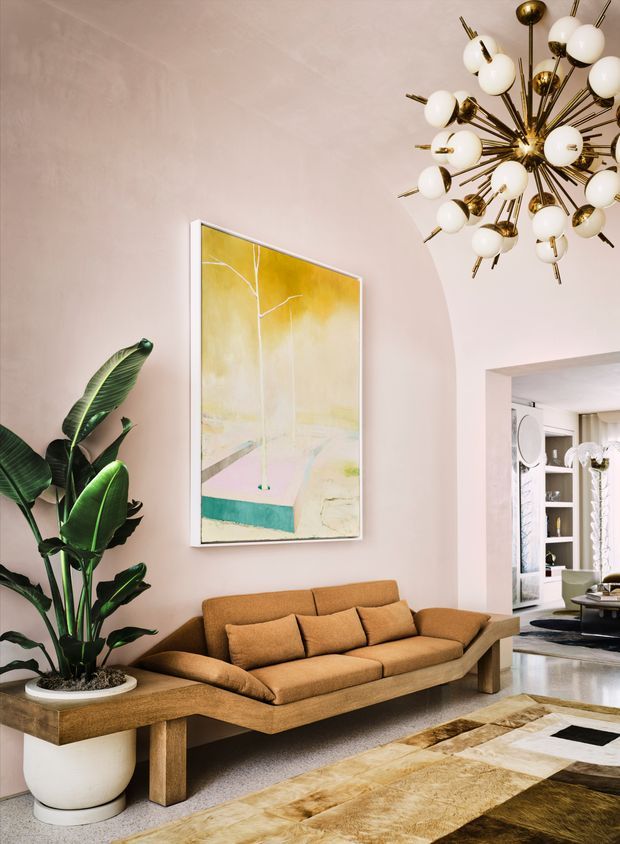 A refined living room with a warm neutral palette, featuring a tan low-profile sofa, substantial timber console or bench, large potted tropical plant, abstract yellow and teal artwork, and a dramatic brass-and-globe Sputnik chandelier. A wide arched opening leads to a bright adjoining space with built-in shelving. The room combines soft plaster walls, timber furnishings, patterned area rugs, and sculptural lighting in an upscale, mid-century-influenced setting.