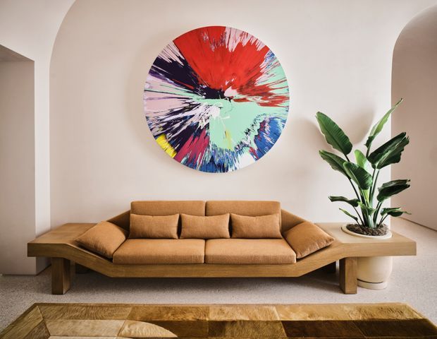 A warm, contemporary living room with a low-profile caramel-brown upholstered sofa set against a softly curved off-white plaster wall. A large, colourful abstract circular artwork forms the focal point above the sofa, while a tall leafy indoor plant sits in a pale ceramic planter to the right. The room features a substantial angular timber sofa base or platform and a geometric timber coffee table in the foreground, with softly integrated arched architectural openings.
